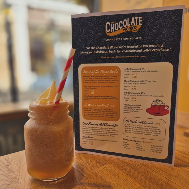 A frosted glass jar of Pop-Star Mocha with a red striped straw, sitting on a wooden table next to the menu card.